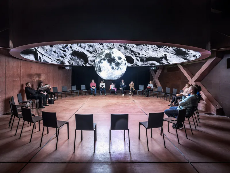 Circular auditorium with corten steel floor and walls showing lunar projection on curved overhead screen with seated audience