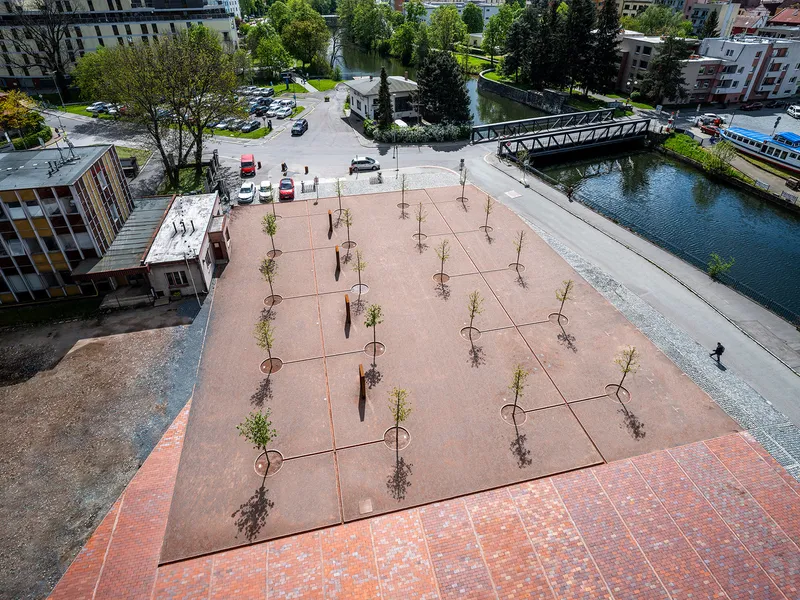 Aerial view of a pink-toned plaza with newly planted trees next to a canal and pedestrian bridge