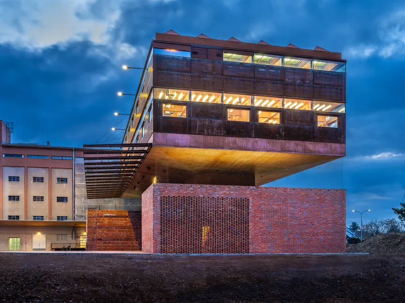 Street view of the cantilevered volume clad in weathered metal panels above a perforated brick base