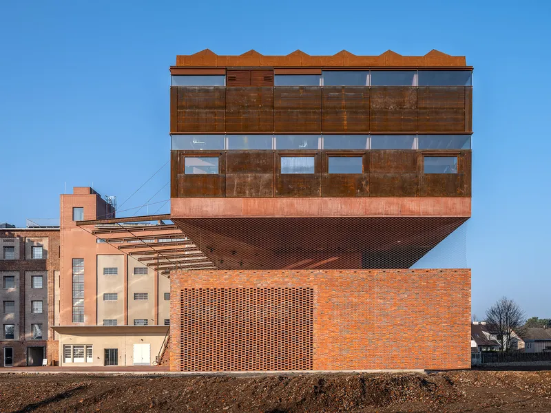 Cantilevered upper volume clad in weathered steel panels above a perforated brick base under clear blue sky
