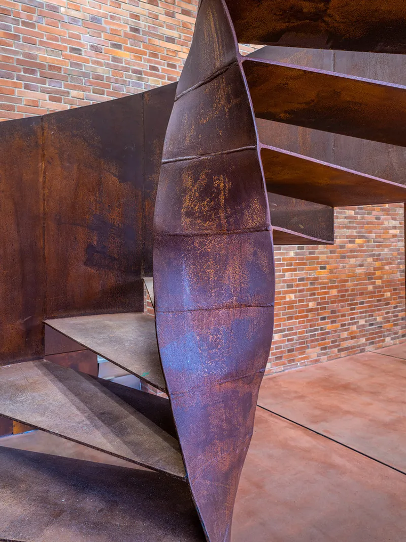 Cantilevered weathered steel staircase with faceted vertical supports against a multi-colored brick wall