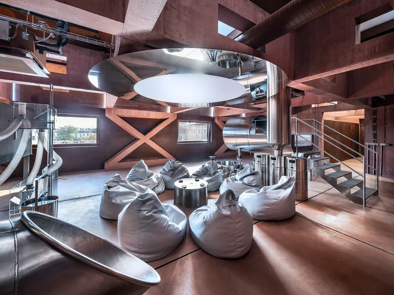 Double-height lounge with exposed brown concrete beams, metallic bean bags, and a circular white pendant fixture overhead