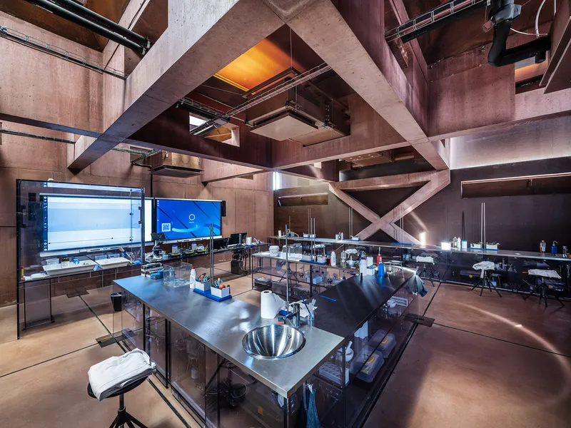 Double-height workshop space with stainless steel workstations beneath crisscrossing concrete beams