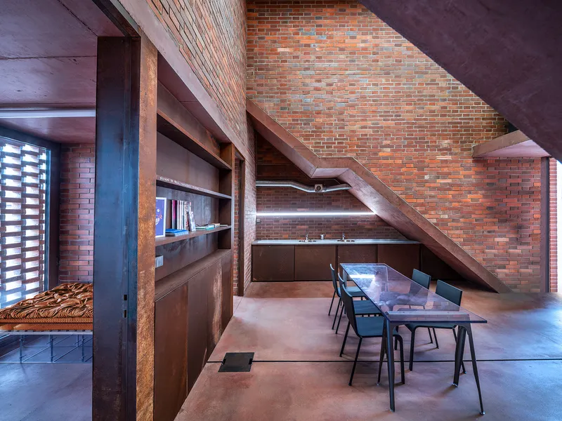 Dining area beneath diagonal concrete staircase with brick wall and built-in shelving