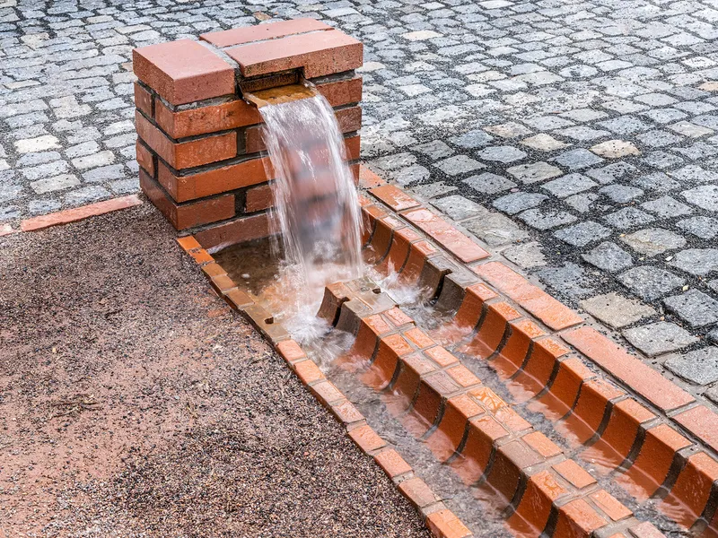 Red brick water fountain with stepped basin flowing over grey cobblestone paving