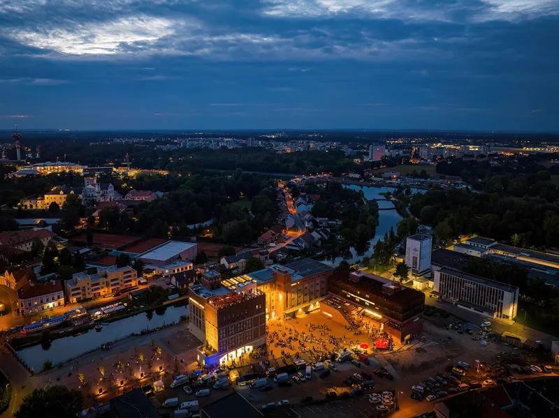Illuminated plaza and brick complex at dusk with river and city lights stretching into the distance