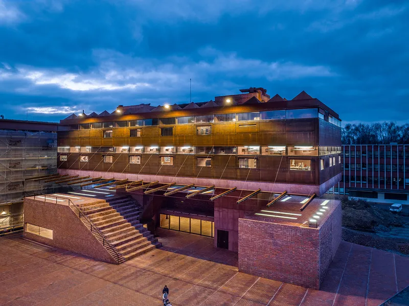 Terraced brick entry plaza with exterior stairs and red facade lit at twilight under cloudy skies