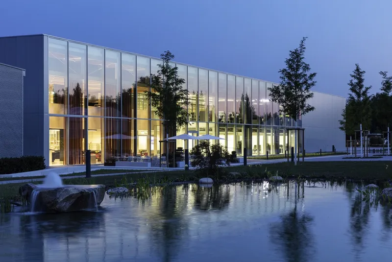 Glass-walled volume beside a reflecting pool with planted evergreens at twilight