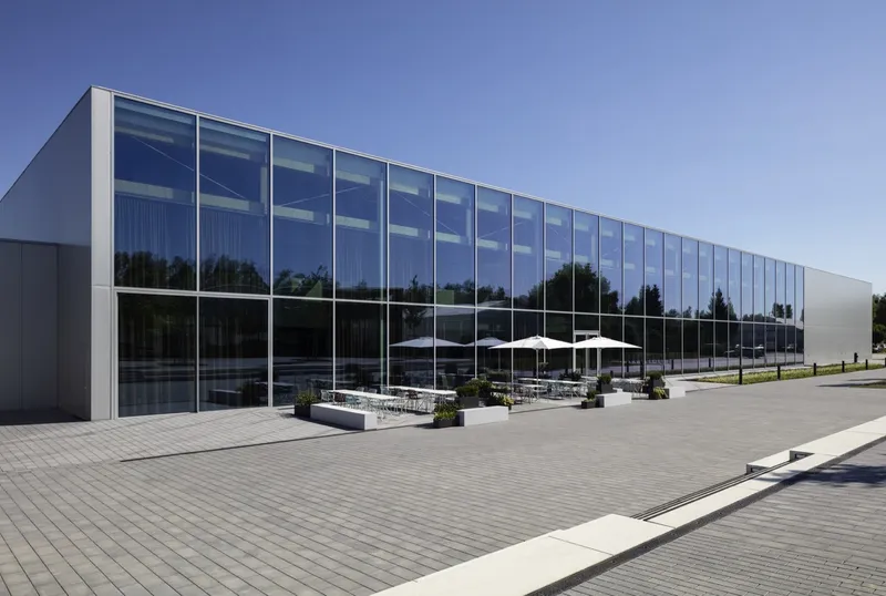Long glass curtain wall reflecting trees and sky above a paved plaza with white umbrellas