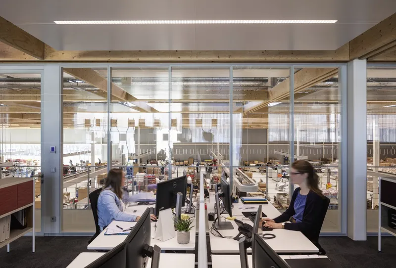 Office desks along glazed partition walls overlooking the production floor with people working at computers