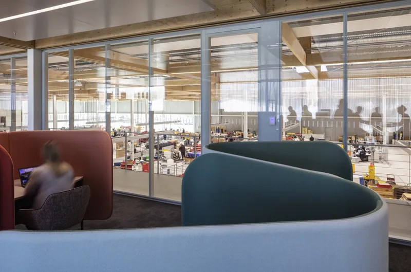 Lounge space with curved upholstered partitions overlooking glass-walled warehouse floor with workers below