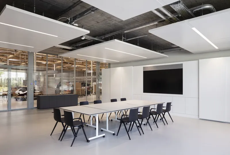 Meeting room with exposed concrete ceiling, suspended acoustic panels and glass partition revealing the workshop beyond