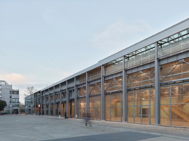 Long facade with exposed steel frame and translucent polycarbonate panels lit from within at dusk