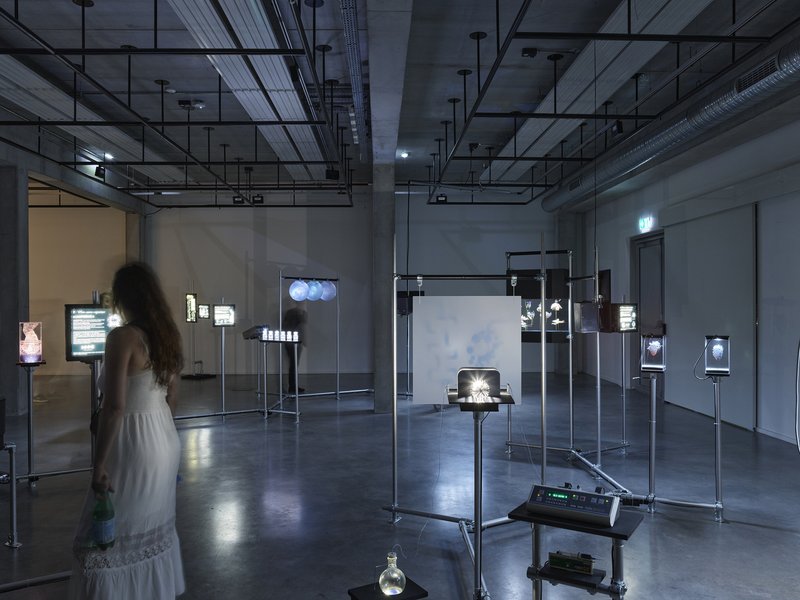 Gallery space with illuminated display screens on metal stands and a visitor in white dress
