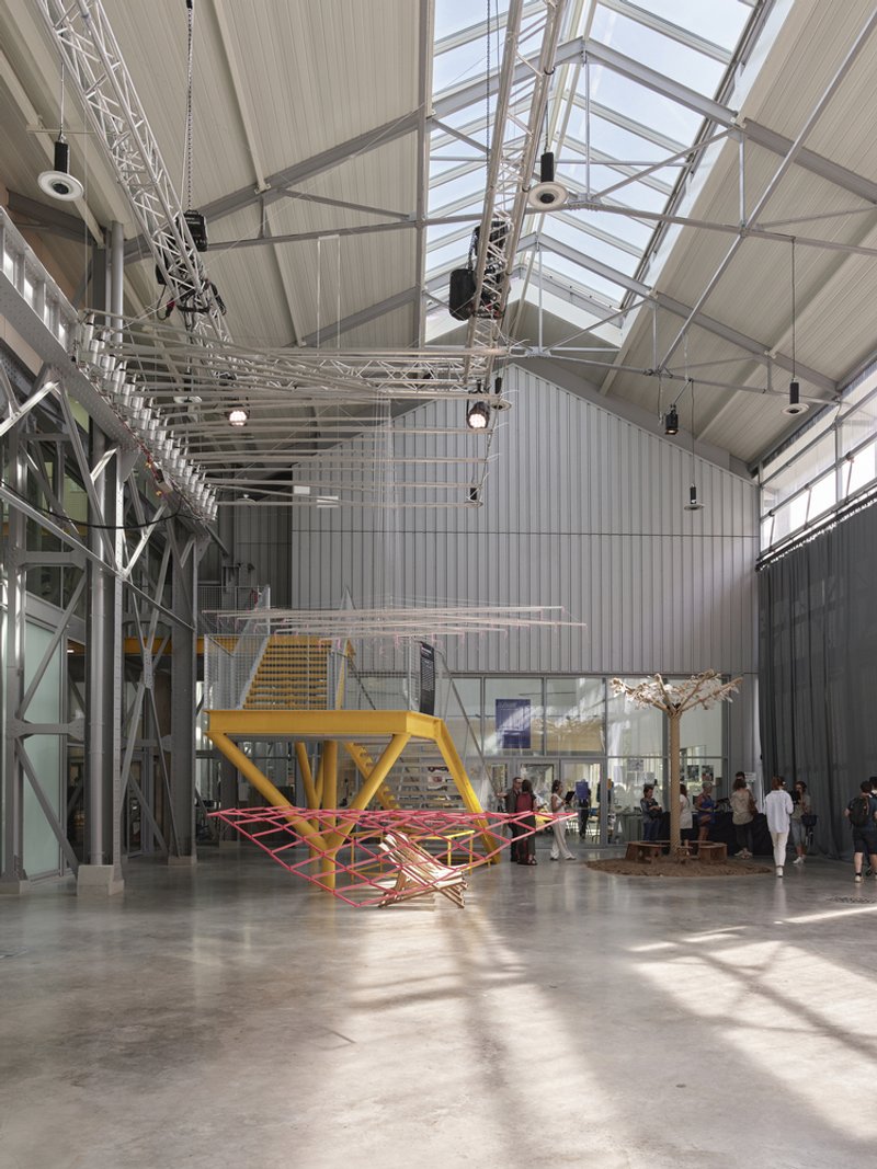 High-ceilinged hall with yellow installation sculpture and suspended lighting truss under clerestory glazing
