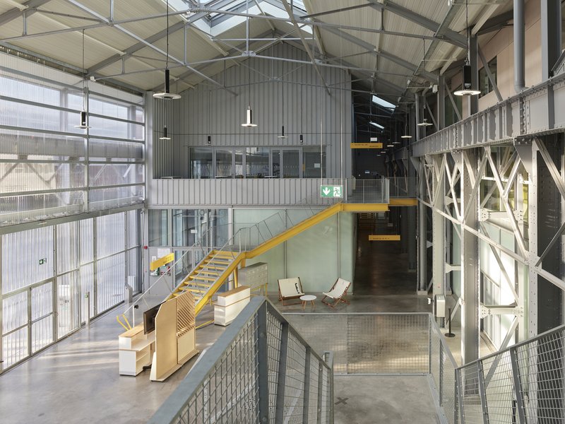 Double-height interior with exposed steel trusses, corrugated metal walls and a yellow staircase descending to the main floor