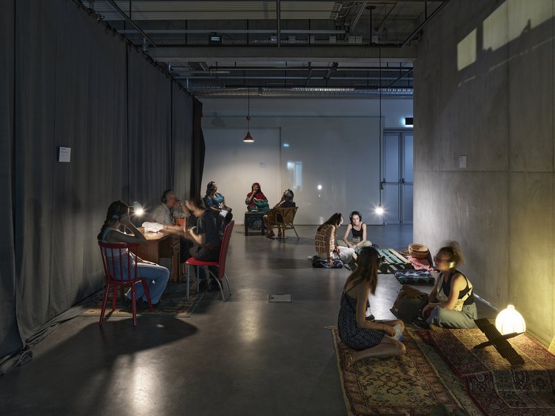Concrete-walled workshop space with exposed ceiling ducts and people gathered around tables and rugs at dusk