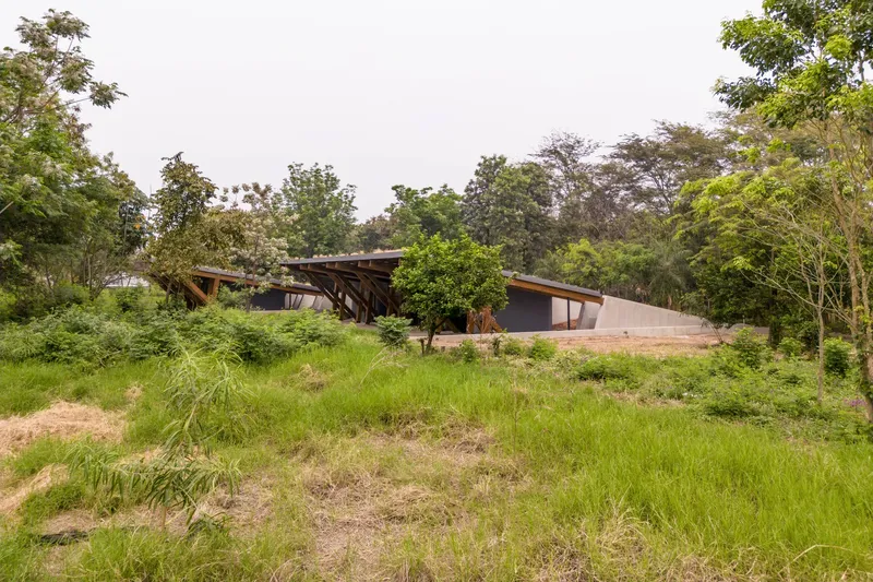 Low pavilion with sloped roofs nestled among tall trees and grassy fields under overcast skies