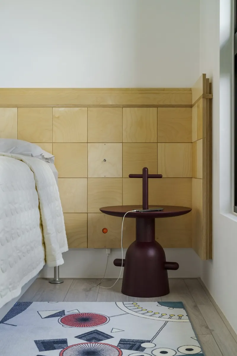 Bedroom corner with light wood paneled wall behind a dark sculptural side table and patterned rug