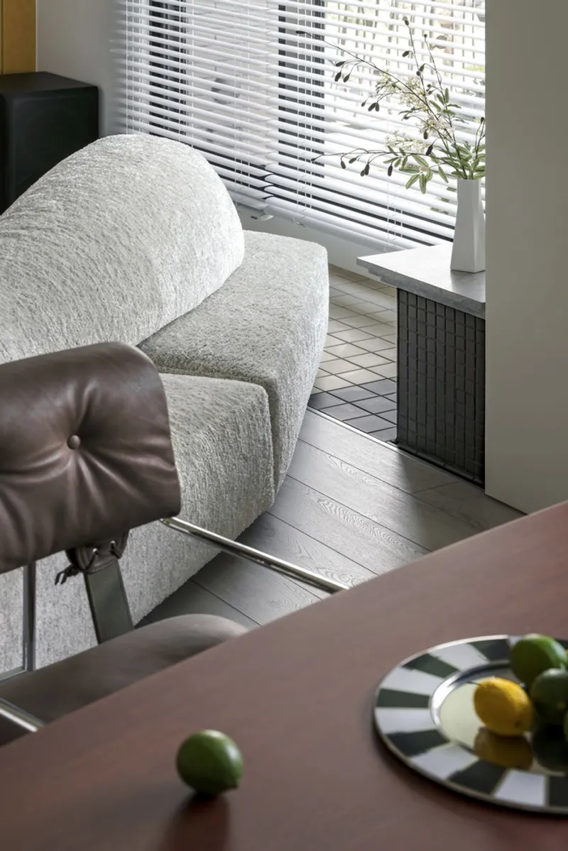 Living area with white horizontal blinds filtering daylight onto pale wood floors and tiled radiator cover