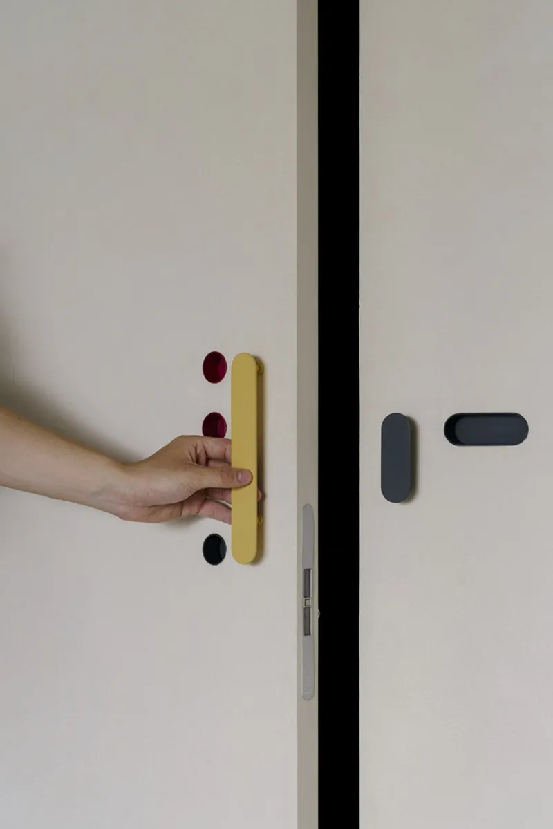 Hand operating yellow door handle with red and black accent buttons on white panel