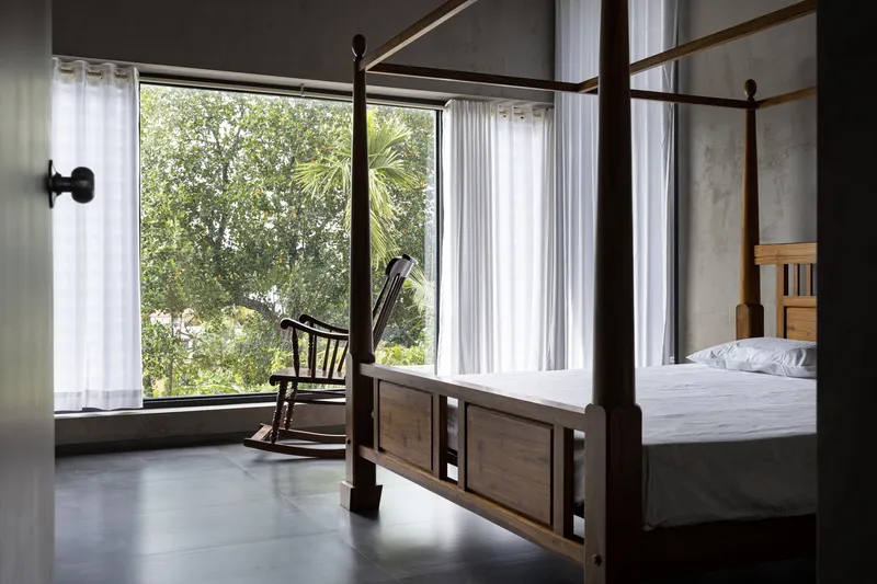 Four-poster bed beside a full-height window framed by white curtains overlooking the garden canopy