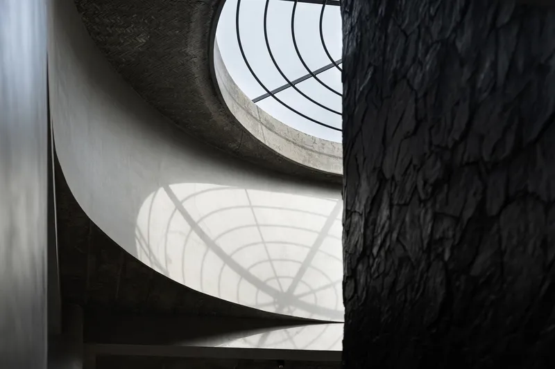 Spiral staircase winding upward past rough black stone wall with natural light filtering through skylight