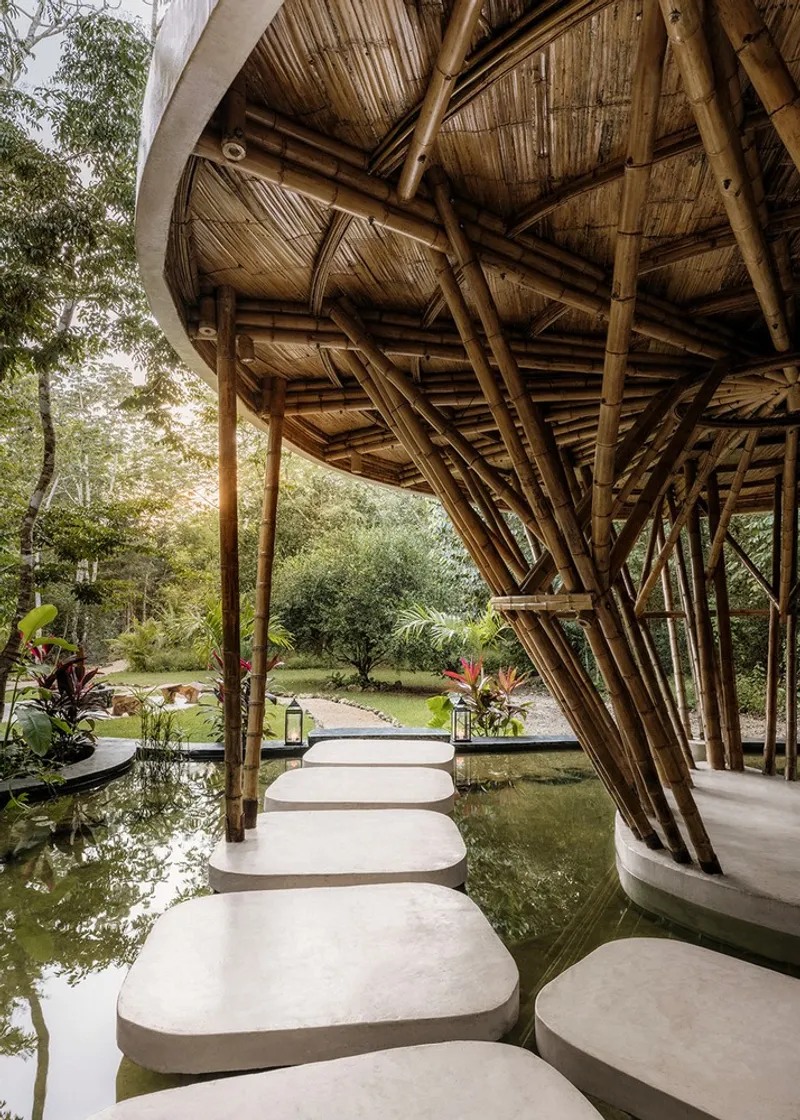 Woven bamboo ceiling structure above concrete stepping stones crossing a shallow reflecting pool toward gardens