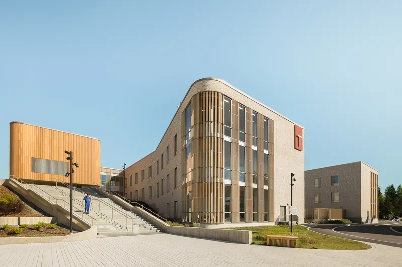 Curved facade corner with timber screen facing the entry plaza and concrete stair on a summer afternoon