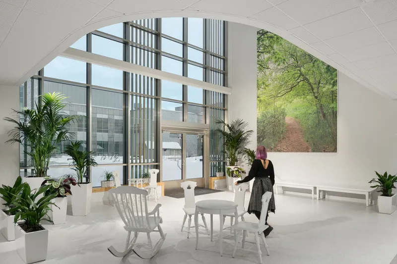 Interior lounge with arched glazing, potted plants, and a seated figure beneath a perforated ceiling