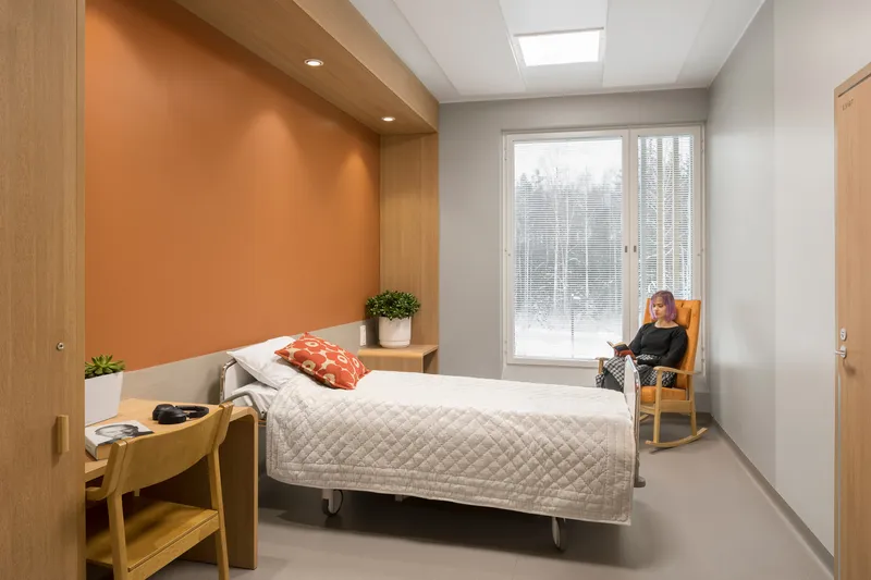 Patient room with timber-framed orange accent wall, a bed, and a woman seated by the window in winter light