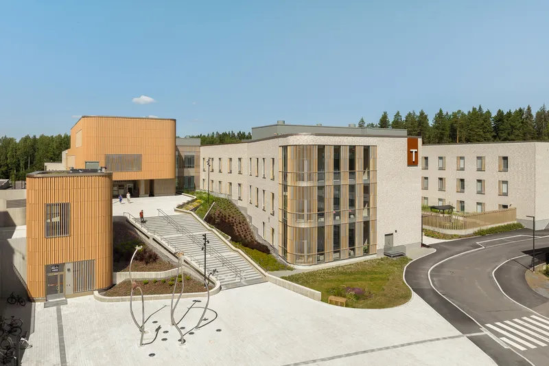 Aerial view of the complex showing vertical timber cladding, cream brick facades, and green roofs beside a forest