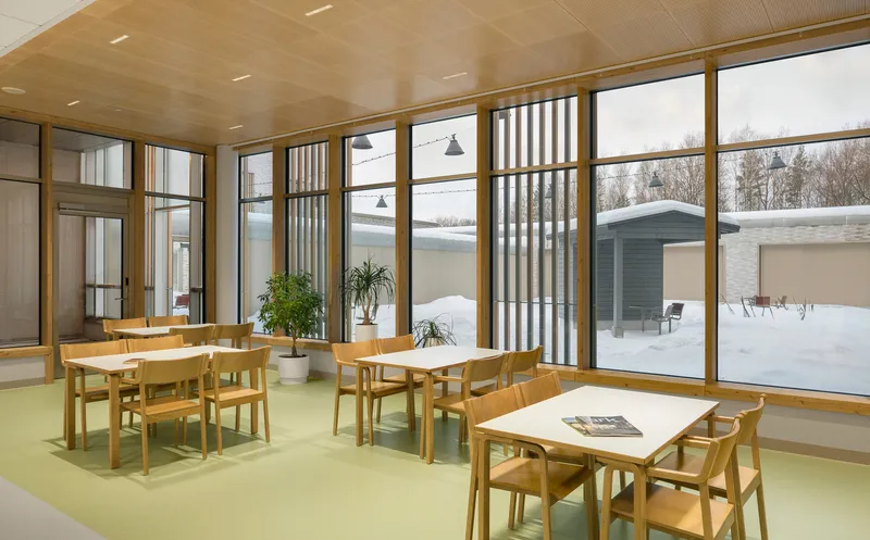 Dining room with timber-framed glazing, pale green flooring, wooden tables, and vertical louvres filtering snowy views