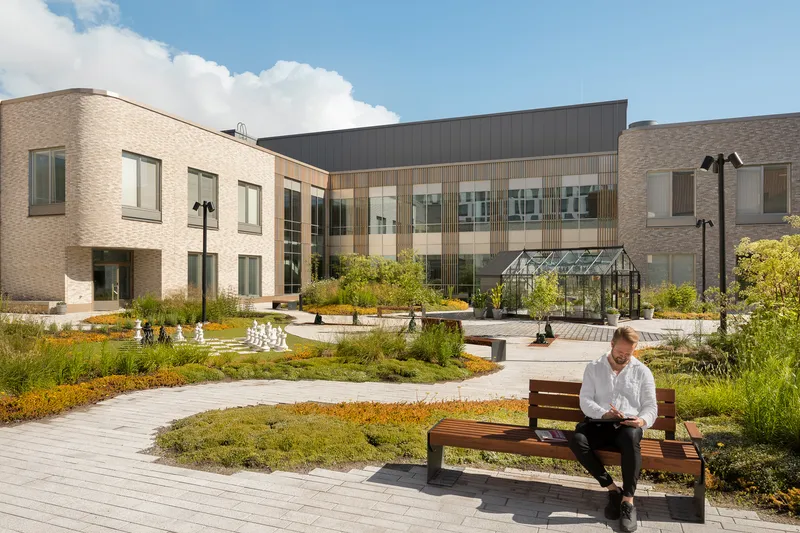 Courtyard with planted beds and timber benches between brick and timber-clad wings under a partly cloudy sky