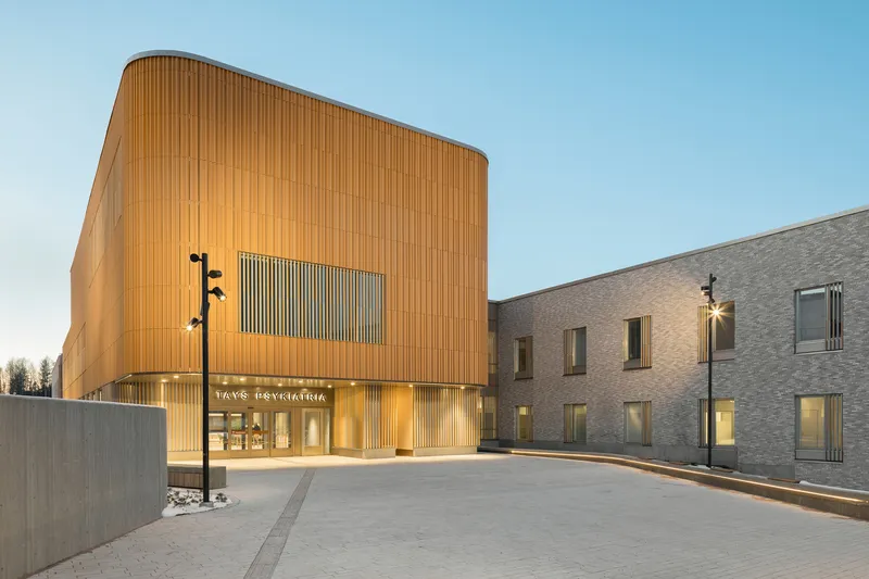 Curved wood-clad facade with vertical slat pattern at dusk next to low brick wings