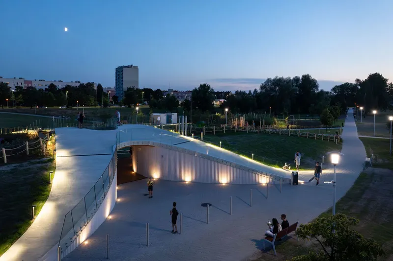 Illuminated ribbon canopy with vertical slat cladding at dusk, casting warm light onto paved pathways