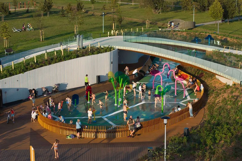 Circular splash pad enclosed by corrugated metal screen walls and timber decking at golden hour