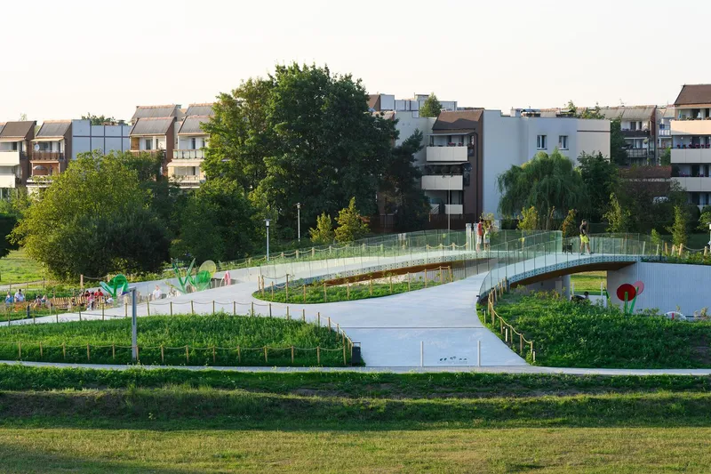 Elevated pedestrian bridge with glass railings spanning between planted beds and residential buildings beyond