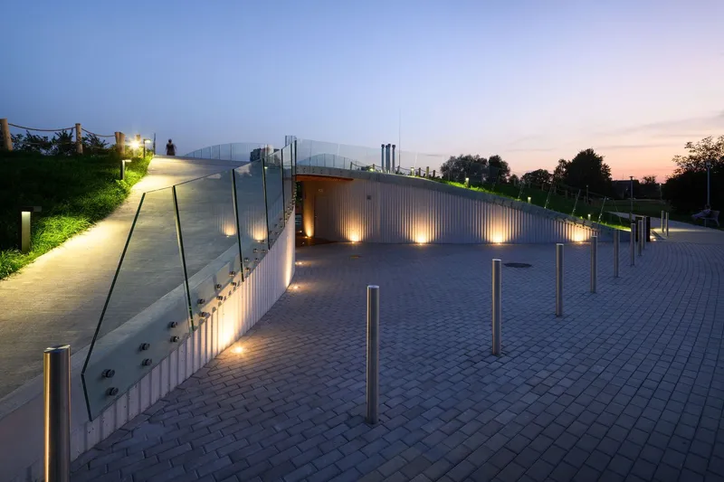 Curved translucent roof entrance with vertical metal cladding and uplighting at dusk