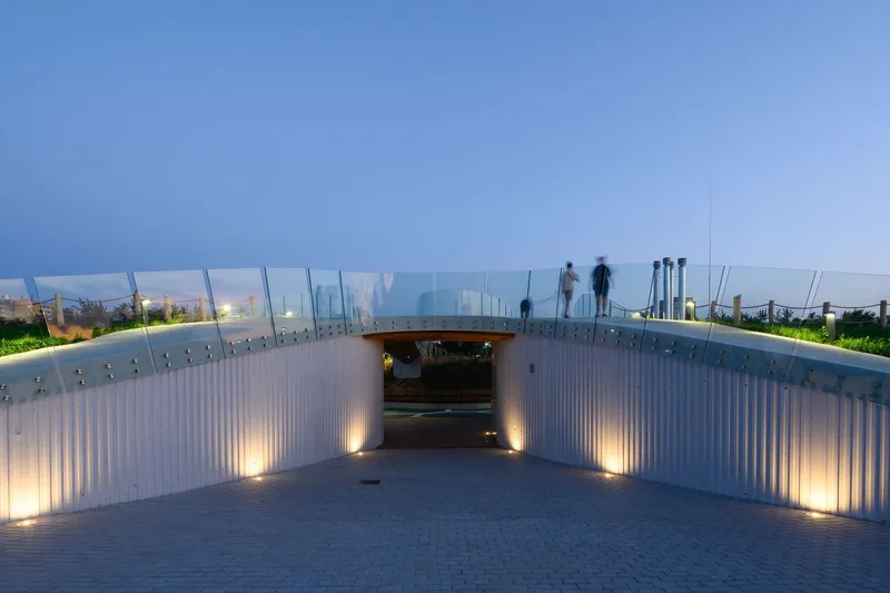 Covered passage with glass balustrade and uplighted corrugated metal walls at twilight with visitors silhouetted above