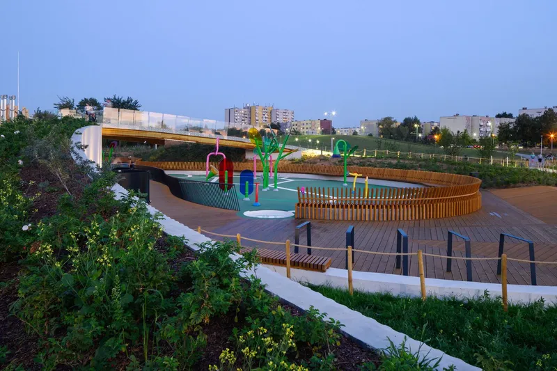 Timber-edged playground and circular deck beside planted slopes at dusk with residential towers in the distance