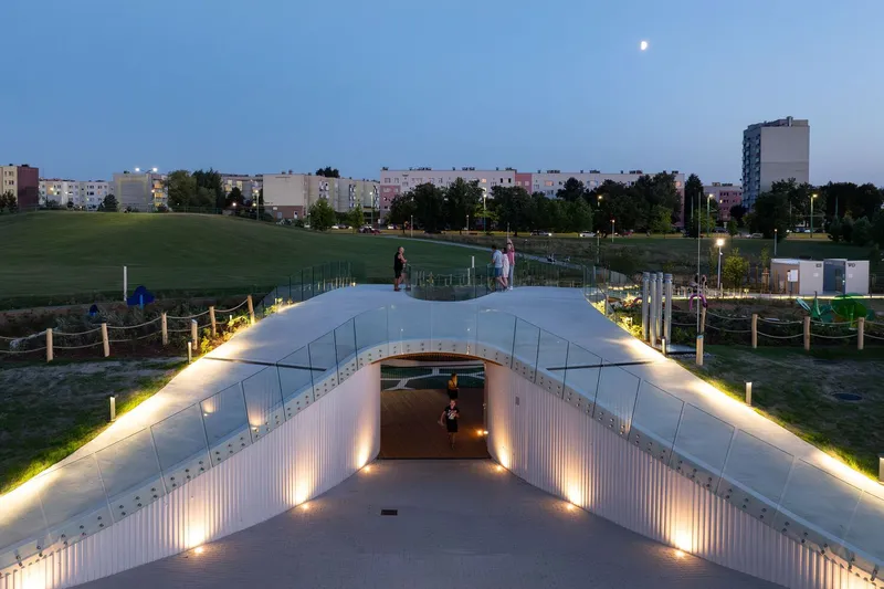Glass-edged ramp descending into illuminated underpass beneath grassy park slope at twilight