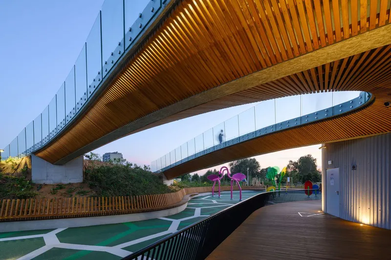 Underside of curved timber-slatted pedestrian bridge spanning over a play area with colorful sculptural elements