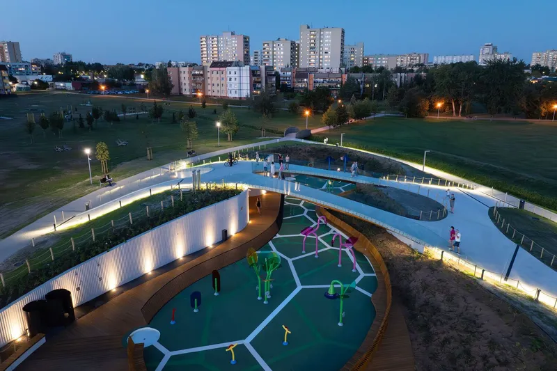 Illuminated curving roof walkway with timber deck wrapping around green play courts in parkland at evening