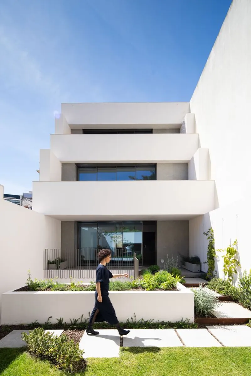 White stepped facade with cantilevered terraces and planted beds as a person walks across the lawn