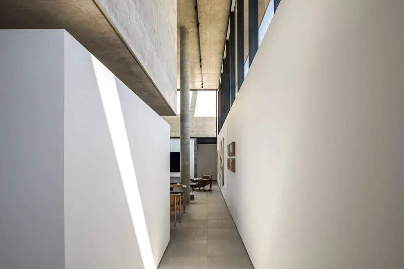 Corridor with white walls and exposed concrete ceiling drawing sunlight toward dining area beyond