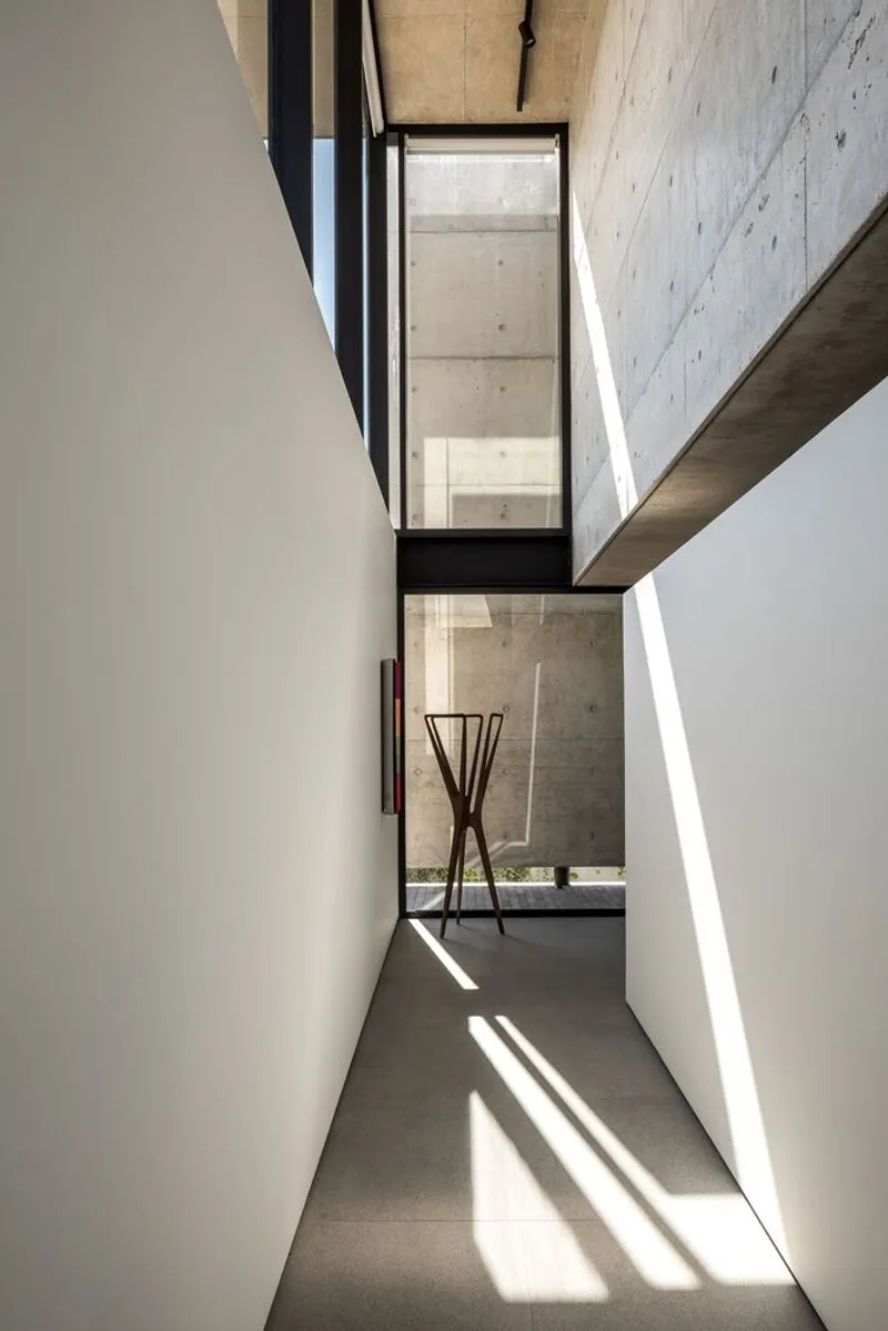 Double-height passageway with concrete walls and black-framed glazed bridge casting angular shadows on floor