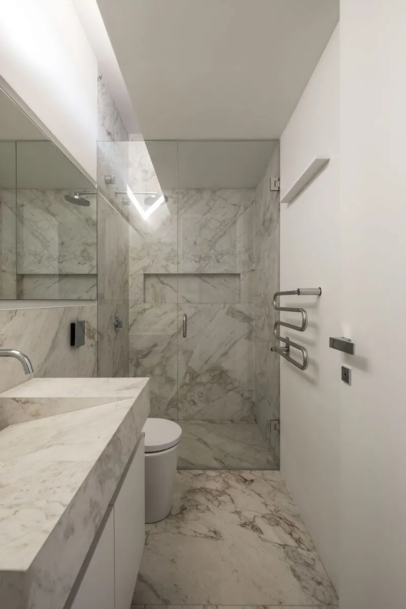Bathroom with white veined marble walls, glass shower enclosure and integrated vanity under linear ceiling light