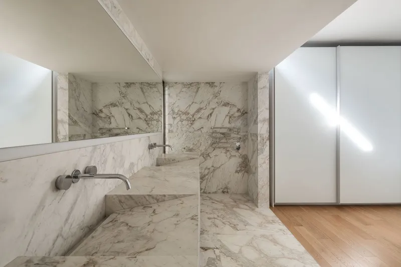 Marble-clad bathtub with wall-mounted faucet and glass partition adjacent to timber flooring