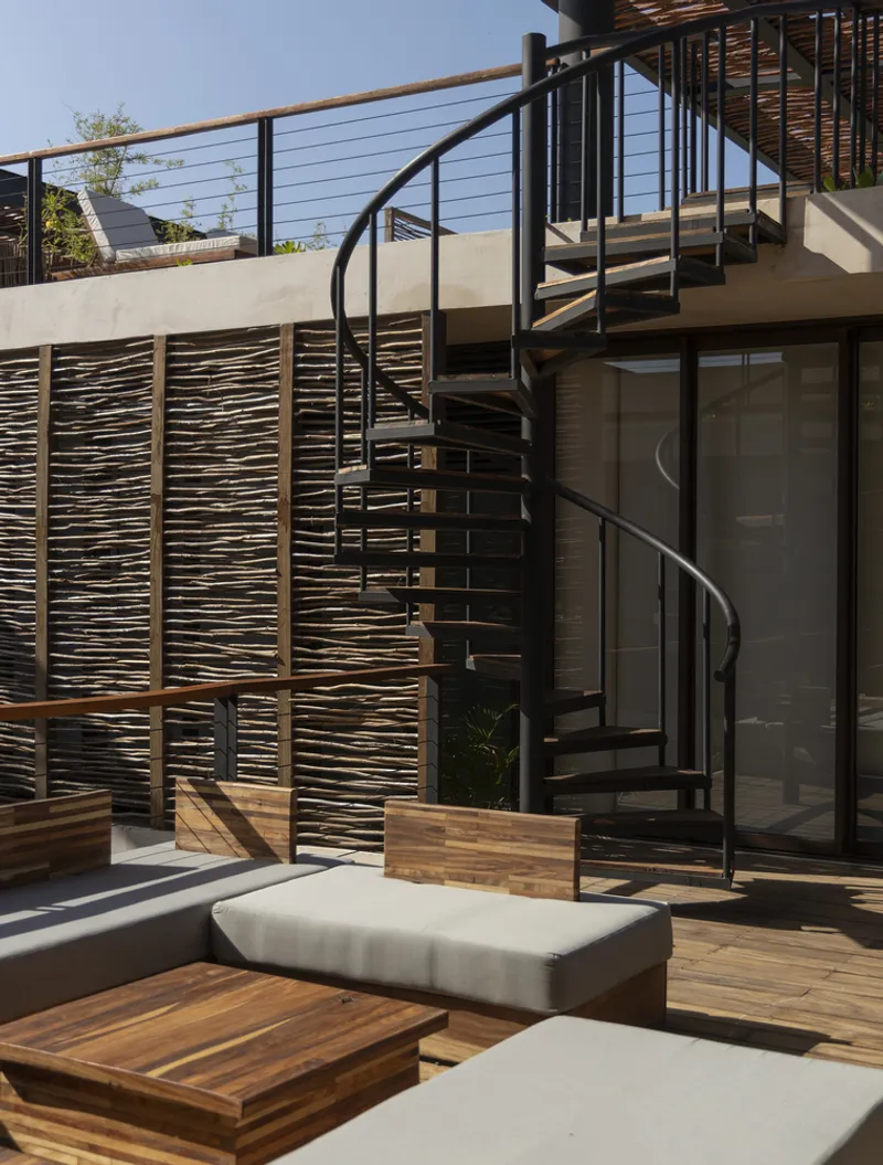 Black spiral stair connecting levels beside woven timber screen and concrete planter boxes in daylight
